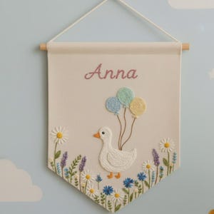 Op de afbeelding: Geborduurde wandhanger met de naam "Anna" in roze schrift. Een witte eend houdt drie kleurrijke ballonnen vast. De onderkant heeft geborduurde bloemen in blauw, wit en paars, wat een grillige kinderkamerdecoratie creëert.