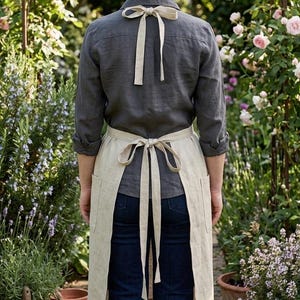May include: A beige linen apron featuring a waist tie and a neck strap, worn over a dark gray long-sleeved button-down shirt and dark blue jeans. The apron includes two side pockets and is tied with a bow at the waist and a bow at the neck.