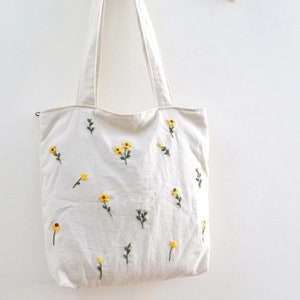 May include: A white canvas tote bag with yellow embroidered flowers. The flowers have green stems and leaves.