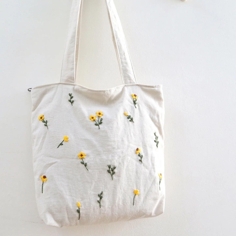 May include: A white canvas tote bag with yellow embroidered flowers. The flowers have green stems and leaves.