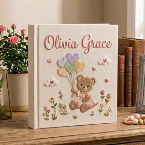 Puede incluir: Libro de recuerdos para beb&eacute;s de color crema con detalles bordados. Presenta el nombre "Olivia Grace" en escritura rosa. Un osito de peluche sosteniendo globos pastel est&aacute; centrado, rodeado de flores y corazones.