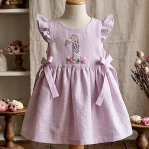 May include: A light purple dress with a floral embroidered number 1 on the front. The dress has ruffled sleeves and bows on the sides. The dress is displayed on a mannequin.
