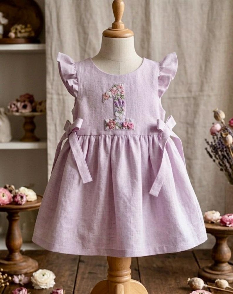 May include: A light purple dress with a floral embroidered number 1 on the front. The dress has ruffled sleeves and bows on the sides. The dress is displayed on a mannequin.