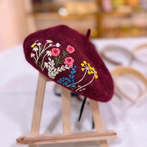May include: A burgundy beret with embroidered floral details. The embroidery features white, pink, yellow, and blue flowers with green stems and leaves. The beret is displayed on a small wooden easel.