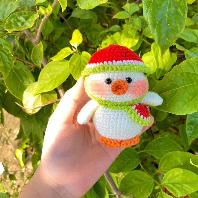 May include: A white crocheted amigurumi toy shaped like a penguin wearing a red and white watermelon hat and a green scarf. The penguin is holding a small watermelon slice.