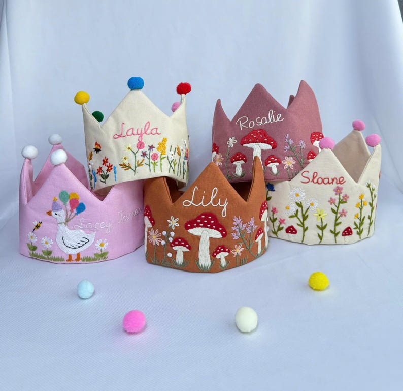 May include: A collection of embroidered fabric crowns in various colors, including pink, tan, and brown. Each crown features unique designs such as flowers, mushrooms, and a goose with balloons. Names like "Layla", "Rosalie", "Sloane", "Lily", and "Lacey" are embroidered on the crowns.