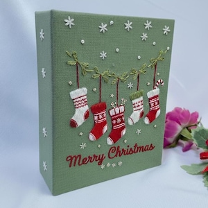 May include: A green rectangular Christmas decoration with embroidered white snowflakes and hanging stockings. The stockings are red and white, and the text "Merry Christmas" is embroidered in red.