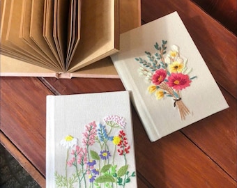 Puede incluir: Dos cuadernos con diseños florales bordados y un diario abierto con páginas marrones. Un cuaderno presenta un ramo de flores rojas, amarillas y rosas. El otro cuaderno tiene un campo de flores coloridas.
