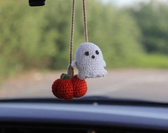 May include: A handmade Halloween car charm featuring a white crocheted ghost and a red crocheted pumpkin. The ghost has black button eyes and is suspended by a brown string. The pumpkin has a green stem. The charm hangs from a car's rearview mirror.