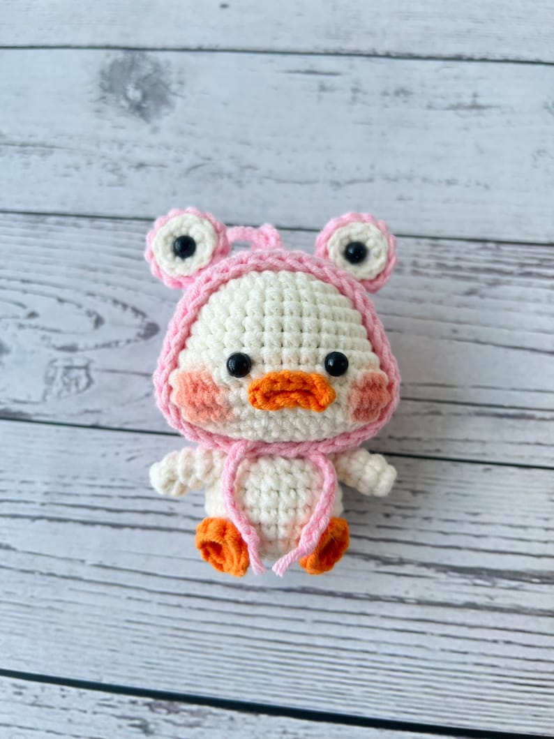 May include: A white crocheted duck wearing a pink frog hat with black eyes. The duck has orange feet and a pink blush on its cheeks.
