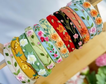 May include: A collection of colorful headbands with embroidered floral designs. The headbands feature various colors, including yellow, green, red, and blue, each adorned with different flower patterns. The headbands are displayed on a wooden stand.