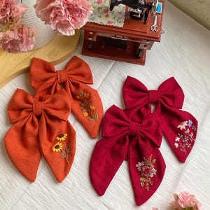 K&ouml;nnte beinhalten: Zwei Paare handgemachter Stoffhaarb&auml;nder mit gestickten Blumenmustern. Ein Paar ist orange mit Sonnenblumen-Stickerei und das andere Paar ist rot mit einer rosa Rose und gr&uuml;nen Bl&auml;ttern Stickerei.