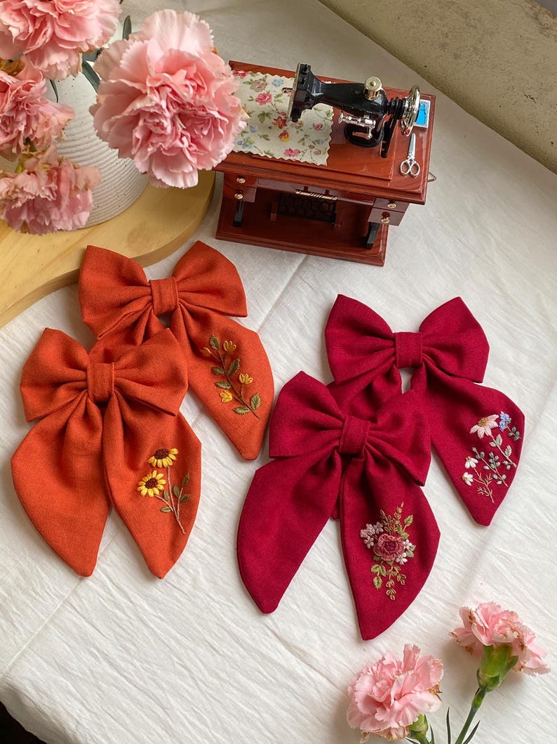 K&ouml;nnte beinhalten: Zwei Paare handgemachter Stoffhaarb&auml;nder mit gestickten Blumenmustern. Ein Paar ist orange mit Sonnenblumen-Stickerei und das andere Paar ist rot mit einer rosa Rose und gr&uuml;nen Bl&auml;ttern Stickerei.