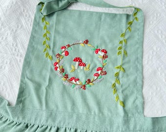 Puede incluir: Un delantal verde con un diseño bordado de setas y hojas. El delantal tiene una falda fruncida y tirantes ajustables.