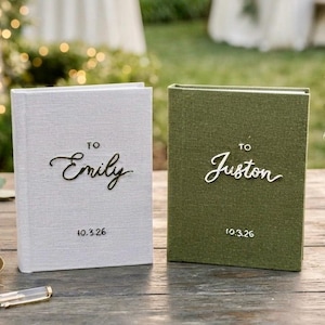 May include: Two hardback books with fabric covers, one light grey and one olive green. The grey book features the text 'TO Emily' and '10.3.26' in black. The green book features the text 'TO Juston' and '10.3.26' in white lettering.