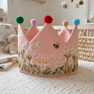 May include: Pink fabric crown embroidered with daisies, green leaves, and the name "Emma". The crown has six points, each topped with a different colored pom-pom. A small embroidered bee is near the name. A charming accessory for a child.