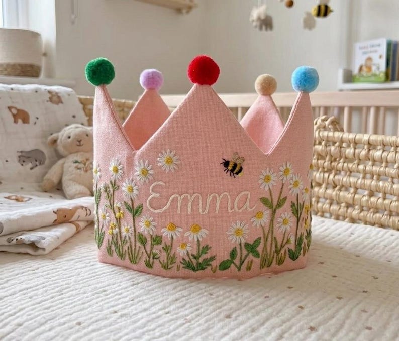 May include: Pink fabric crown embroidered with daisies, green leaves, and the name "Emma". The crown has six points, each topped with a different colored pom-pom. A small embroidered bee is near the name. A charming accessory for a child.