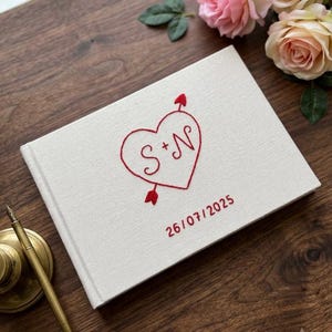 Puede incluir: Un libro de visitas cubierto de tela blanca con detalles bordados en rojo. La portada muestra un corazón atravesado por una flecha con las letras S + N en su interior, y la fecha 26/07/2025 bordada en rojo debajo del diseño del corazón.