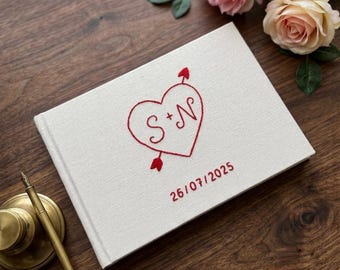 May include: A white fabric-covered guest book featuring red embroidered details. The cover displays a heart pierced by an arrow with the letters S + N inside, and the date 26/07/2025 embroidered in red below the heart design.