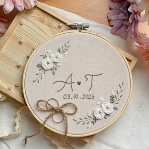 Op de afbeelding: Geborduurde ringhouder met initialen "A T" en de datum 03.10.2025. De beige stof is geplaatst in een houten borduurring, versierd met witte bloemenborduurwerk en een touwstrik.