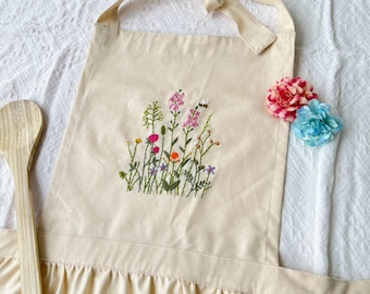 May include: A beige cotton apron with a floral embroidery design. The embroidery features a variety of colorful flowers and a bee. The apron has a ruffled bottom and adjustable straps.