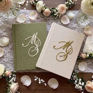 Pode incluir: Dois livros de capa dura com revestimento em tecido, um em verde-oliva e outro em creme. O livro verde apresenta iniciais bordadas em cursivo branco, enquanto o livro creme apresenta iniciais bordadas em cursivo dourado. Acabamento texturizado.