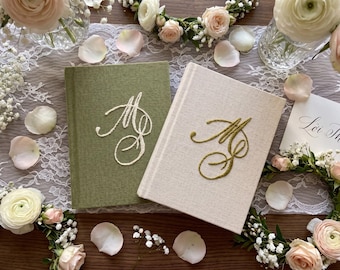 May include: Two hardcover books with fabric covers, one in olive green and one in cream. The olive book features white embroidered cursive initials, while the cream book features gold embroidered cursive initials. Both books have a textured finish.
