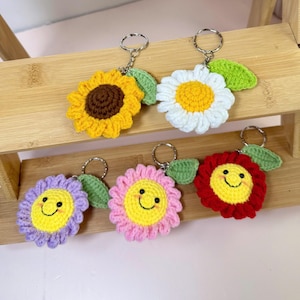 Può includere: Una collezione di portachiavi a forma di fiore all'uncinetto fatti a mano in vari colori, tra cui giallo, bianco, viola, rosa e rosso. Ogni portachiavi presenta un volto sorridente e un accento di foglia verde, attaccato a un portachiavi argentato.