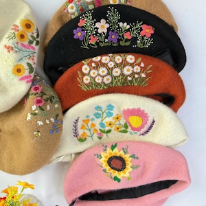 May include: A collection of berets in various colors, each adorned with intricate floral embroidery. The berets feature designs such as sunflowers, daisies, and other colorful blooms, showcasing a handcrafted aesthetic. The berets are in shades of pink, black, brown, and cream.