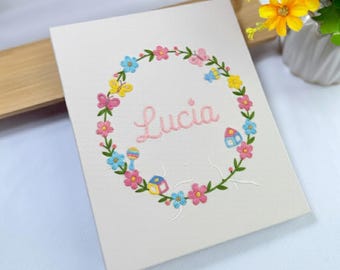 May include: Embroidered canvas art featuring the name "Lucia" in pink script, surrounded by a floral wreath with butterflies, flowers, and a small house. The wreath includes pink, yellow, and blue elements. The background is off-white.