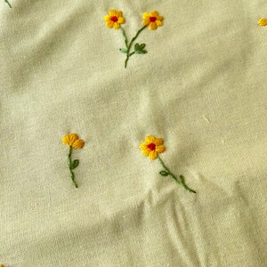 May include: A close-up of a light beige fabric with yellow embroidered flowers. The flowers have red centers and green stems.