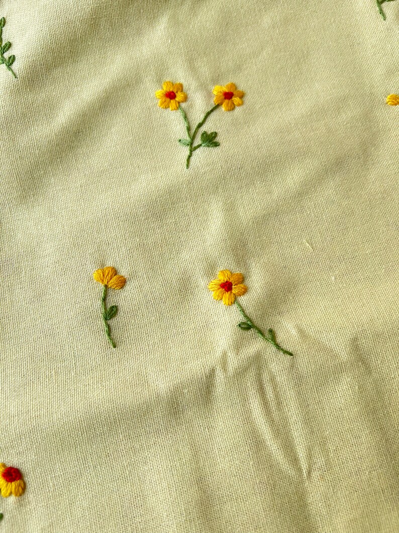 May include: A close-up of a light beige fabric with yellow embroidered flowers. The flowers have red centers and green stems.