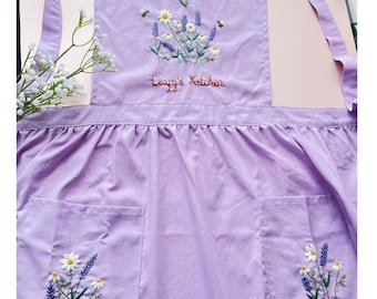 May include: A lavender apron with embroidered floral designs, including daisies, lavender, and bees. The apron has two pockets and the text "Leapy's Kitchen" embroidered on the front. The apron has long straps for tying around the waist and neck.