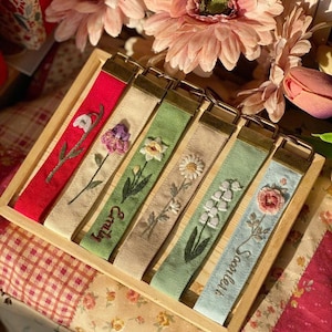 May include: Six embroidered fabric keychains are displayed in a wooden tray. Each keychain has a different colour and floral design, with names such as "Emily" and "Scarlett" embroidered on some. The keychains have gold-coloured metal hardware.