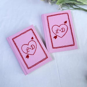 Puede incluir: Dos cuadernos rosas bordados con diseños de corazón y flecha rojos. Cada cuaderno presenta las iniciales "L+D" dentro del corazón, rodeadas por un borde rojo decorativo. Los cuadernos están sobre una superficie blanca.