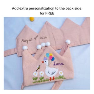 May include: Pink fabric crown with embroidered details. The crown features a white goose holding balloons, the name "Luna", and the date "August 9th 2024". Additional text reads "Add extra personalization to the back side for FREE".