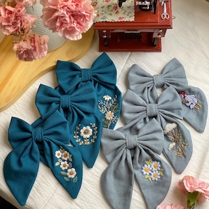 K&ouml;nnte beinhalten: F&uuml;nf handgefertigte Stoffhaarb&auml;nder mit gestickten Blumenmustern. Zwei Schleifen sind blaugr&uuml;n mit wei&szlig;en Blumen und drei Schleifen sind hellblau mit wei&szlig;en und violetten Blumen.