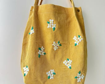 May include: A yellow corduroy tote bag with white embroidered flowers. The bag has two straps and a zip closure.