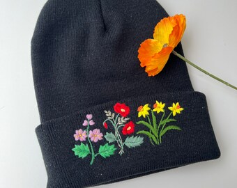 Puede incluir: Gorro negro con flores bordadas en tonos rojos, rosas, amarillos y verdes. El gorro está doblado hacia abajo y una sola flor naranja descansa en la parte superior.