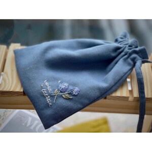 May include: A light blue drawstring bag with a floral embroidery design. The embroidery features two small purple flowers with green stems and leaves. The text "voila la vie" is embroidered in white below the flowers.