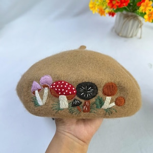 May include: A tan wool beret with an embroidered mushroom design. The embroidery features various colorful mushrooms, including red, purple, and brown, with green foliage. The beret is held in a hand.