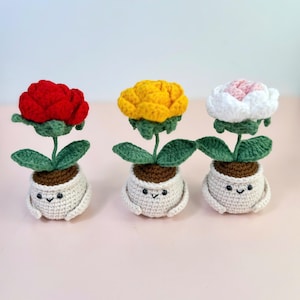 May include: Three crocheted flower pot dolls. Each doll has a different colored flower: red, yellow, and white with pink accents. The pots are white with smiling faces and small arms. The flowers have green leaves.