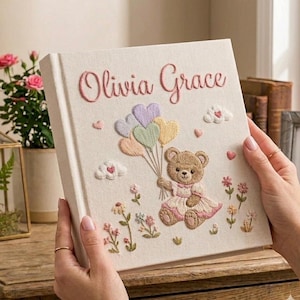 Puede incluir: Un libro de recuerdos para beb&eacute;s de color crema con el nombre "Olivia Grace" bordado en rosa. La portada presenta un osito de peluche sosteniendo globos en forma de coraz&oacute;n, bordados en colores pastel, con flores y nubes.
