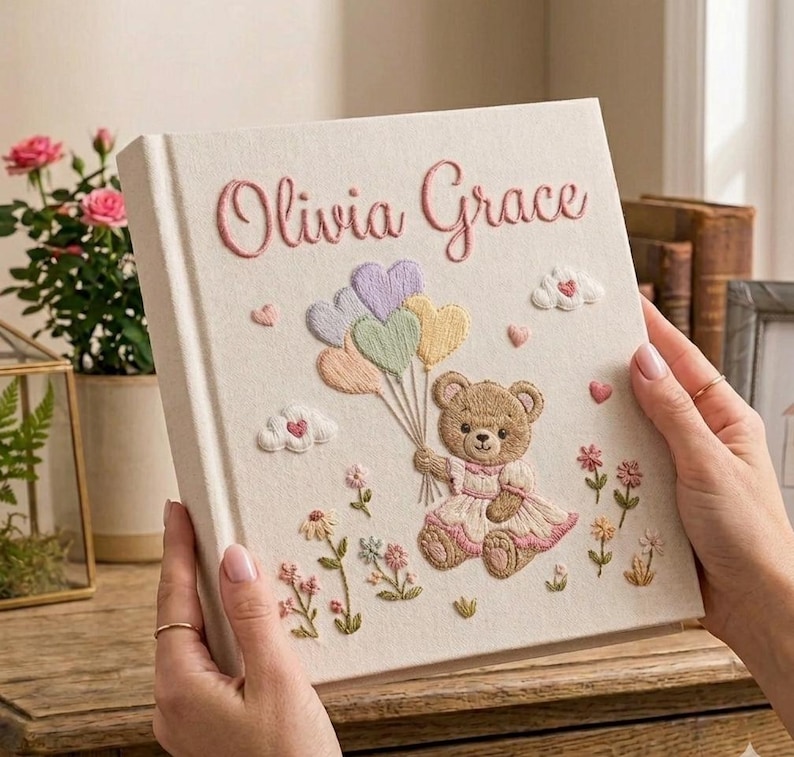 Puede incluir: Un libro de recuerdos para beb&eacute;s de color crema con el nombre "Olivia Grace" bordado en rosa. La portada presenta un osito de peluche sosteniendo globos en forma de coraz&oacute;n, bordados en colores pastel, con flores y nubes.
