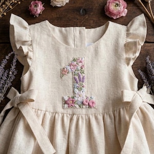 May include: A cream-colored linen dress with a floral embroidered number "1" on the front. The dress features ruffled sleeves and side bows. The embroidery includes pink, purple, and white flowers.