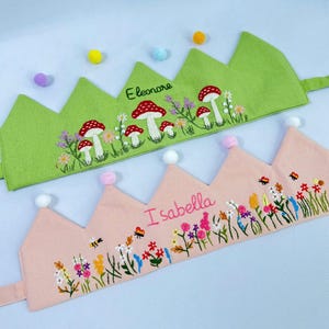 May include: Two embroidered fabric crowns. One is light green with red and white mushroom and floral embroidery, the name "Eleonore" is stitched on the crown. The other is pink with floral and bee embroidery, and the name "Isabella".