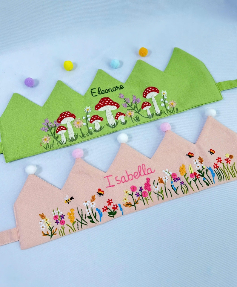 May include: Two embroidered fabric crowns. One is light green with red and white mushroom and floral embroidery, the name "Eleonore" is stitched on the crown. The other is pink with floral and bee embroidery, and the name "Isabella".