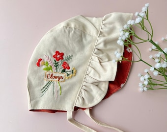 May include: A beige baby bonnet with a red floral embroidery and the name "Elowyn". The bonnet has a ruffled edge and a red interior. It has tie strings for securing. The bonnet is likely made of linen or a similar fabric.