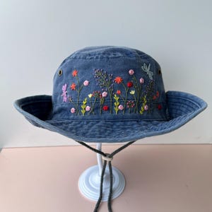 Puede incluir: Un sombrero de pescador de mezclilla azul con flores bordadas y una libélula en tonos de rosa, naranja, amarillo y verde.