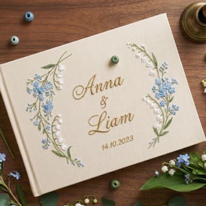 Puede incluir: Un libro de visitas de boda color crema con diseños florales bordados en azul y blanco. Los nombres "Anna & Liam" están bordados en dorado, junto con la fecha "14.10.2023". El libro está sobre una superficie de madera con elementos decorativos.
